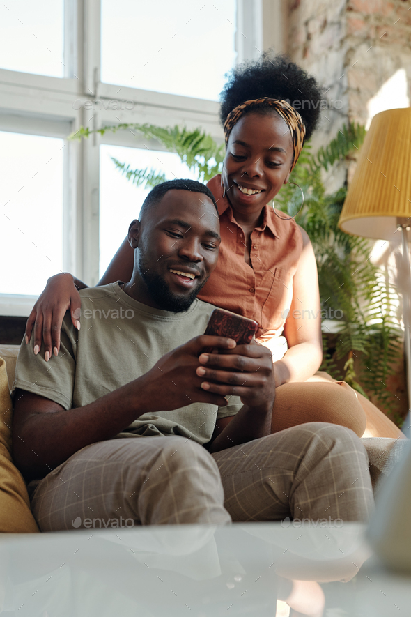 Joyful young African couple in casualwear looking at smartphone screen with smiles