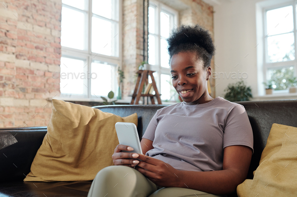 Restful young African woman with smartphone sitting on black leather couch and watching funny video