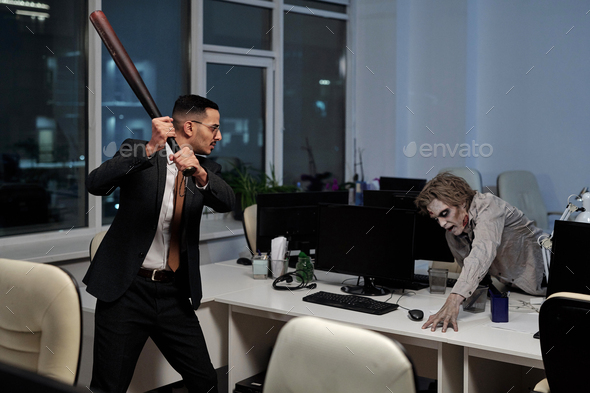 Young angry businessman with baseball bat going to hit male zombie trying to get upon desk