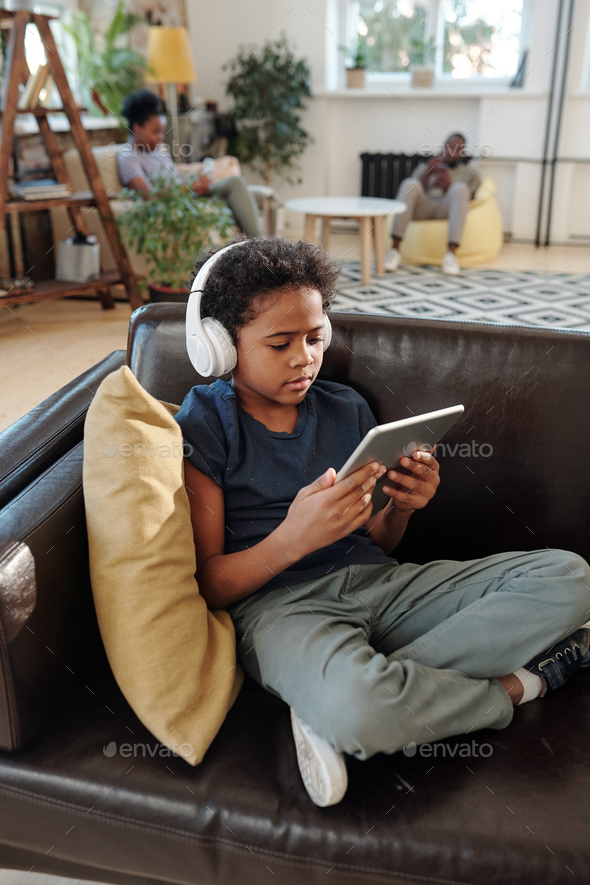 Adorable little boy with headphones sitting on black leather couch and watching online video