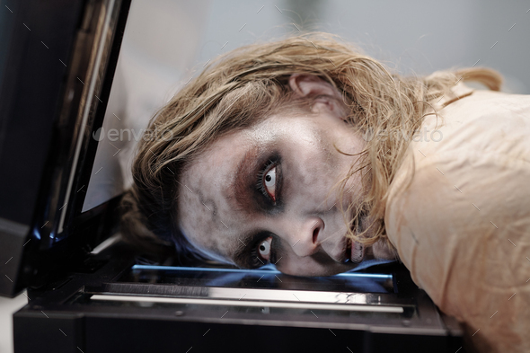 Woman with open eyes and zombie greasepaint on face keeping her head on screen of xerox machine