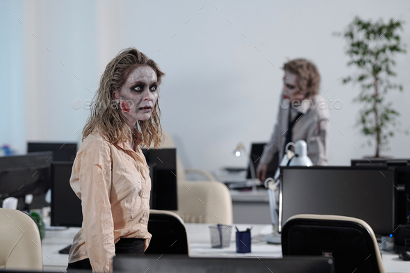 Young zombie businesswoman walking between desks with computer monitors against dead male colleague