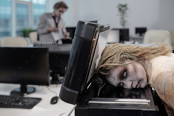 Young spooky businesswoman with zombie greasepaint on face keeping head on screen of xerox machine