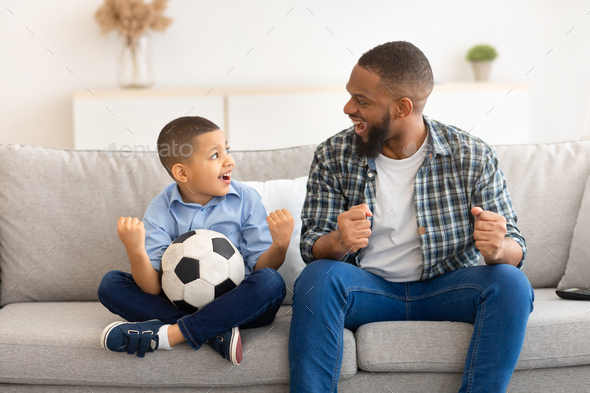 African Dad And Son Shaking Fists Watching Soccer At Home