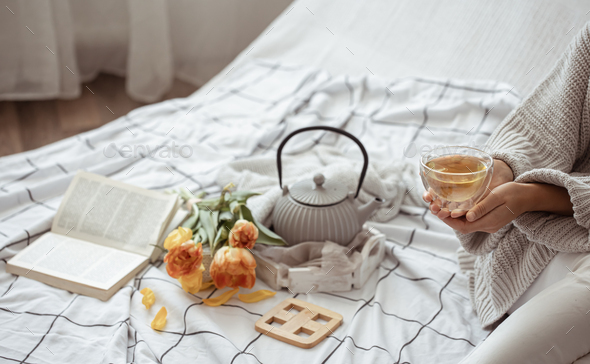 Spring home composition in bed with tea in female hands, a teapot, a book and a bouquet of flowers.