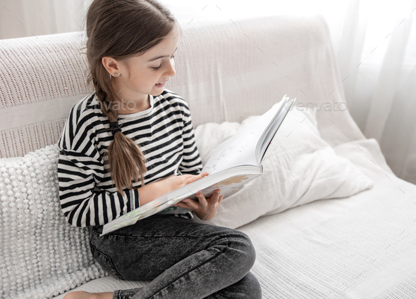 Little cheerful girl reads a book while sitting on the couch at home.