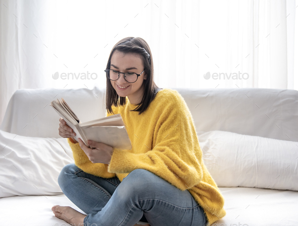 A cute girl in glasses and a yellow sweater is sitting on the sofa with a book.