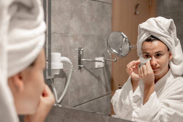 A young woman in a robe and with a towel on her head is putting patches under her eyes.
