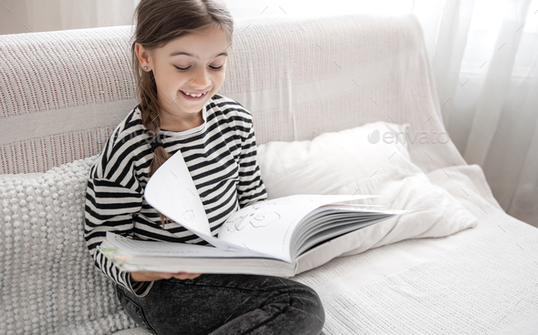 Little cheerful girl reads a book while sitting on the couch at home.