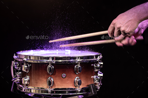 Drum sticks hitting snare drum with splashing water on black background under studio lighting.