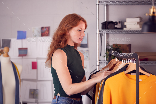 Female Owner Of Fashion Business Checking Clothing Samples On Rail In Studio