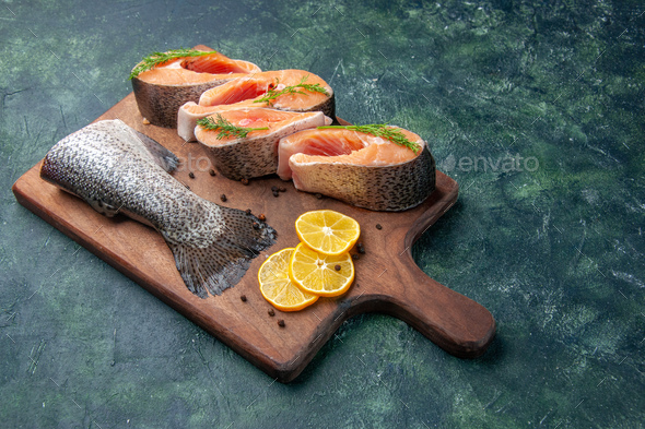 Front view of fresh raw fishes lemon slices greens pepper on wooden cutting board on dark mix