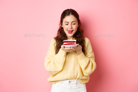 Young slim girl lick her lips from temptation, looking at delicious piece of cake with yearning to