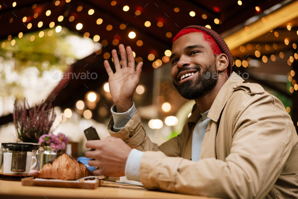 African american man waving and holding cellphone in cafe outdoors