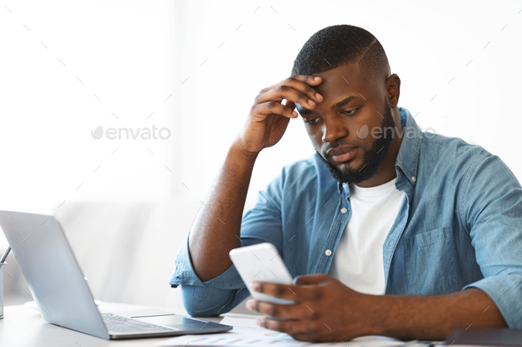 Concerned black freelancer guy looking at smartphone while working at home office - Stock Photo - Images