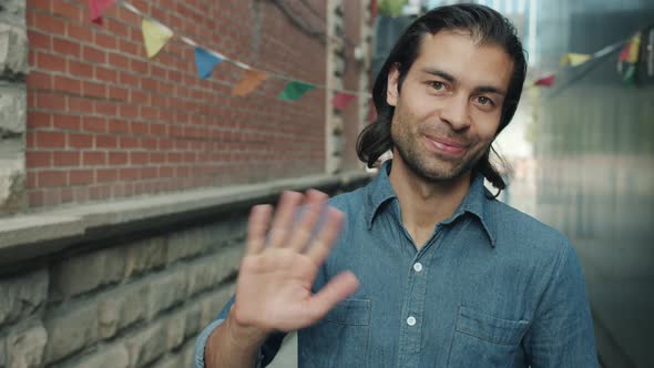 Smiling Mixed Race Guy Waving Hand Greeting Looking at Camera Outdoors with City in Background alt