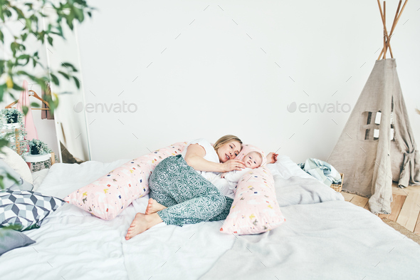 A beautiful young mother with long blonde hair and a daughter of 2-3 months are resting on the bed