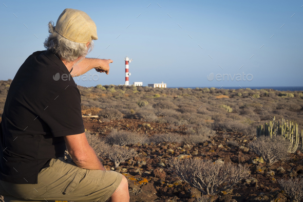 Adult senior man with cap indicates the light house on the cliff, tropical area with volcanic rocks - Stock Photo - Images