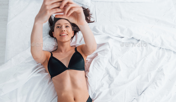 Slender woman in black underwear lying down on bed indoors in room
