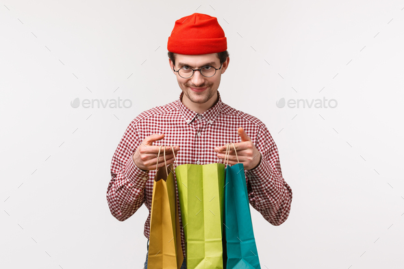 Funny and excited young caucasian hipster guy in red beanie, glasses, open shopping bags and looking
