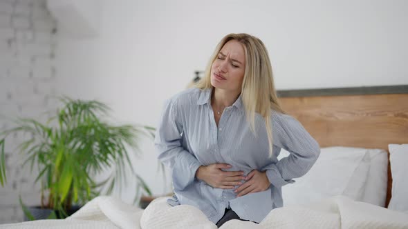 Portrait of Unwell Young Caucasian Woman with Stomach Ache Sitting on Bed at Home alt