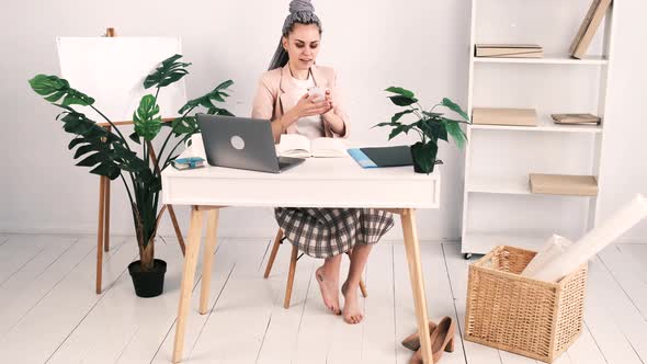 Attractive Young Barefoot Caucasian Woman in a Pink Jacket Sits in an Office at a Table with an Open alt