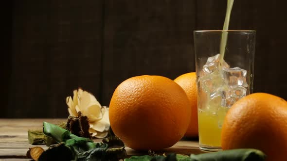Pour Orange Juice Into A Glass With Ice And Three Oranges Next To It On A Table With Flowers