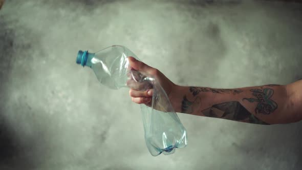 Man's Hand with Tattoos Squeezing Plastic Bottle on Grey Background. Plastic Recycling Concept.