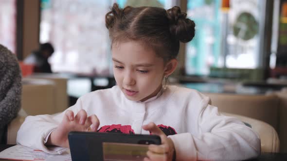 Little Girl Playing on Smartphone the Game Online Sitting at a Table in a Cafe