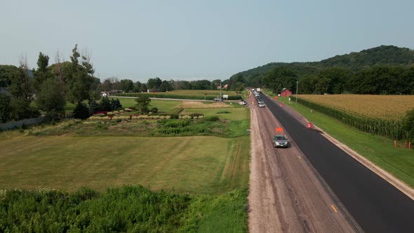 Drone view over rural highway being repaired with new asphalt. Lush green farm field. Blue sky.