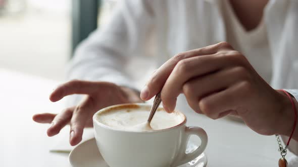 A Hand Stirs A Cup Of Coffee With A Spoon, Cappuccino Coffee In A White Cup Stirring The Coffee Foam alt