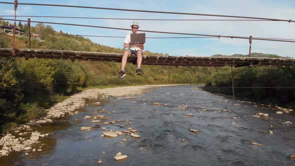 Aerial Drone View of Young Man in White Shirt Sitting on Wooden Bridge Over Mountains River and