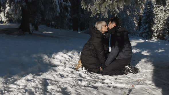 Beautiful Couple Enjoying a Winter Day in the Forest sitting in Snow Holding Hands and Kissing alt