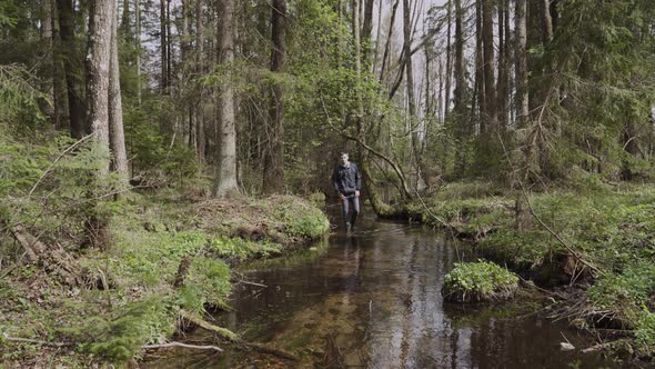 Young Man in Rubber Boots Walks Along the Bed of a Clear Stream in the Forest alt