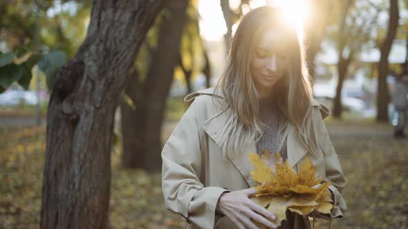 Thoughtful Woman with Pile of Golden Leaves in Hands in Sunny Autumn Park alt