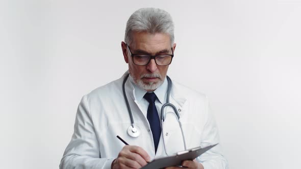 Old Doctor in White Coat with Stethoscope Makes Record in Tablet and Looks at the Camera on White alt