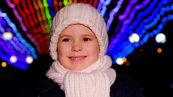 A Handsome Child in a White Hat Smiles Standing Under Christmas Garlands in the Evening alt