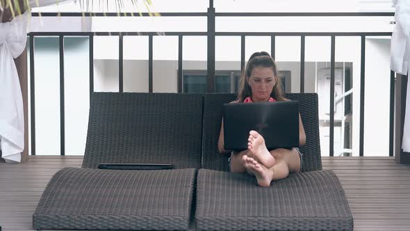 Barefoot Girl Sits with Laptop on Couch in Tropical Hotel alt