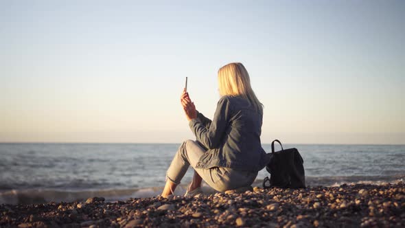 a woman sits on the seashore and writes a sea sunset on her phone. alt