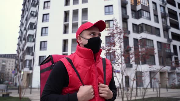 Delivery Guy Wearing Red Uniform and Black Mask While Walking Along Modern Buildings Down City