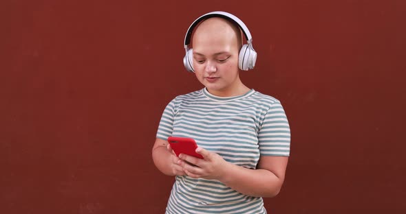Young bald girl using mobile phone app to listening music playlist while wearing wireless headphones alt