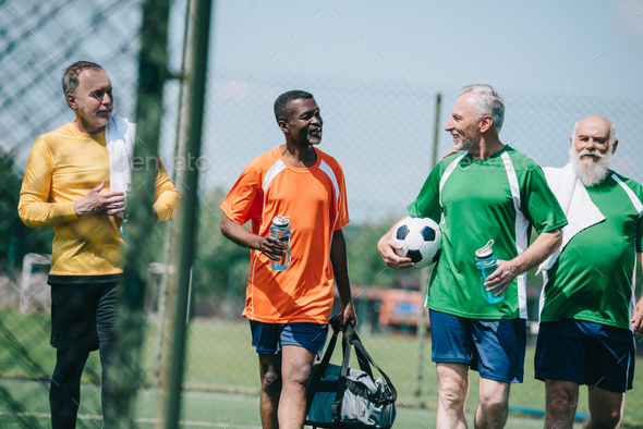 group of interracial elderly sportsmen with sportive water bottles walking on football field