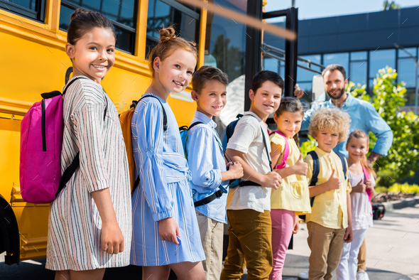 queue of happy pupils standing near school bus with teacher