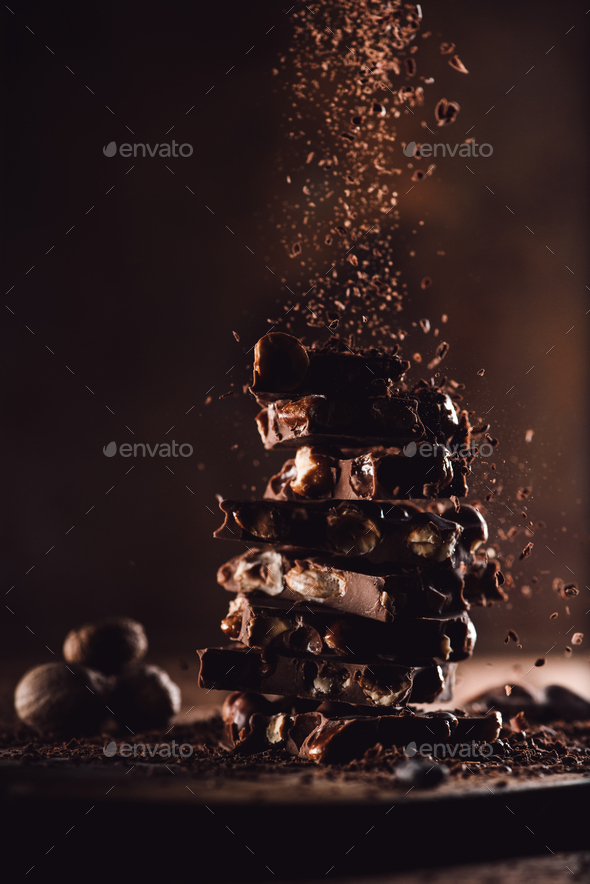 closeup view of nutmegs and grated chocolate falling on stack of chocolate pieces on wooden table