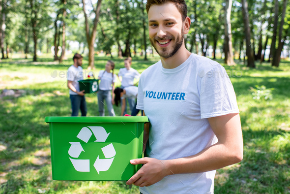 young smiling volunteer with green recycling box in park with friends on background