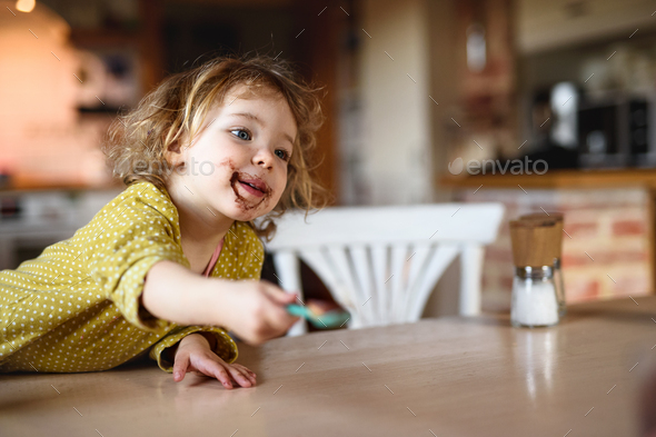 Small girl with dirty mouth indoors in kitchen at home, eating pudding