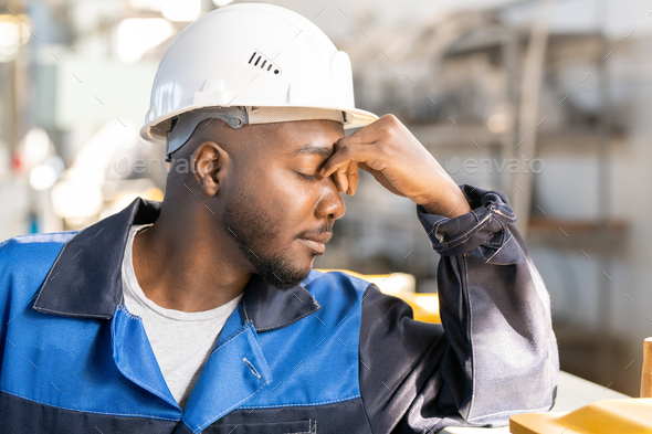 Young tired factory worker of African ethnicity in workwear and hardhat touching his forehead