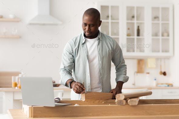 African american man assembling furniture, watching online instructions on laptop computer at home