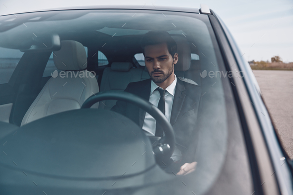 Hurrying to get things done. Handsome young man in full suit looking straight while driving a car