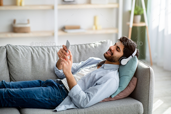 Free time. Relaxed arab guy listening to music in wireless headphones, lying on comfy couch
